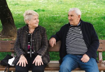 Elder couple laughing while sitting on park bench. Senior husband and wife enjoying retirement on sunny day outdoors