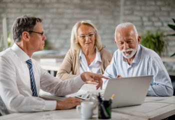 Senior couple using laptop with their financial advisor during a meeting int he office. Focus is on senior man.
