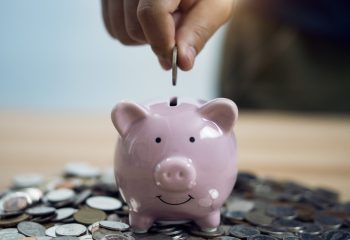 Person Putting Coin In Piggy Bank At Table