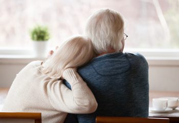 Rear view at senior grey haired loving caring family couple embracing relaxing at home together enjoying peaceful morning breakfast looking at window view thinking of future feeling calm nostalgic