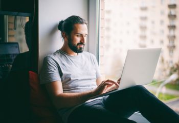 Handsome attractive young dark-haired man working on laptop at home, sitting on windowsill with city view
