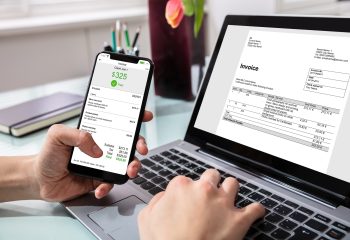 Close-up Of Businessperson Using Laptop While Paying Invoice On Mobilephone In Office