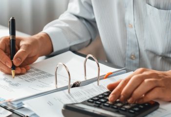 Corporate auditor calculating budget with calculator on his office desk. Dedicated accountant professional of accounting business company analyzing financial document to forecast income. Insight