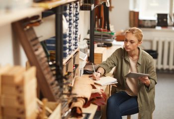 Horizontal high angle portrait of serious young woman sitting at table in craft worshop holding tablet PC and making notes in notebook