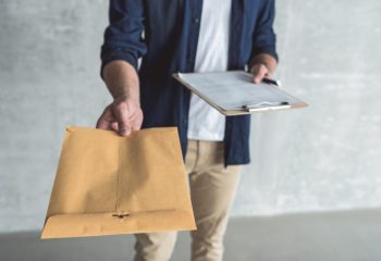 This is for you. Close-up of envelope in hand of diligent professional courier. He is standing with package and personal contract against gray wall background. Selective focus